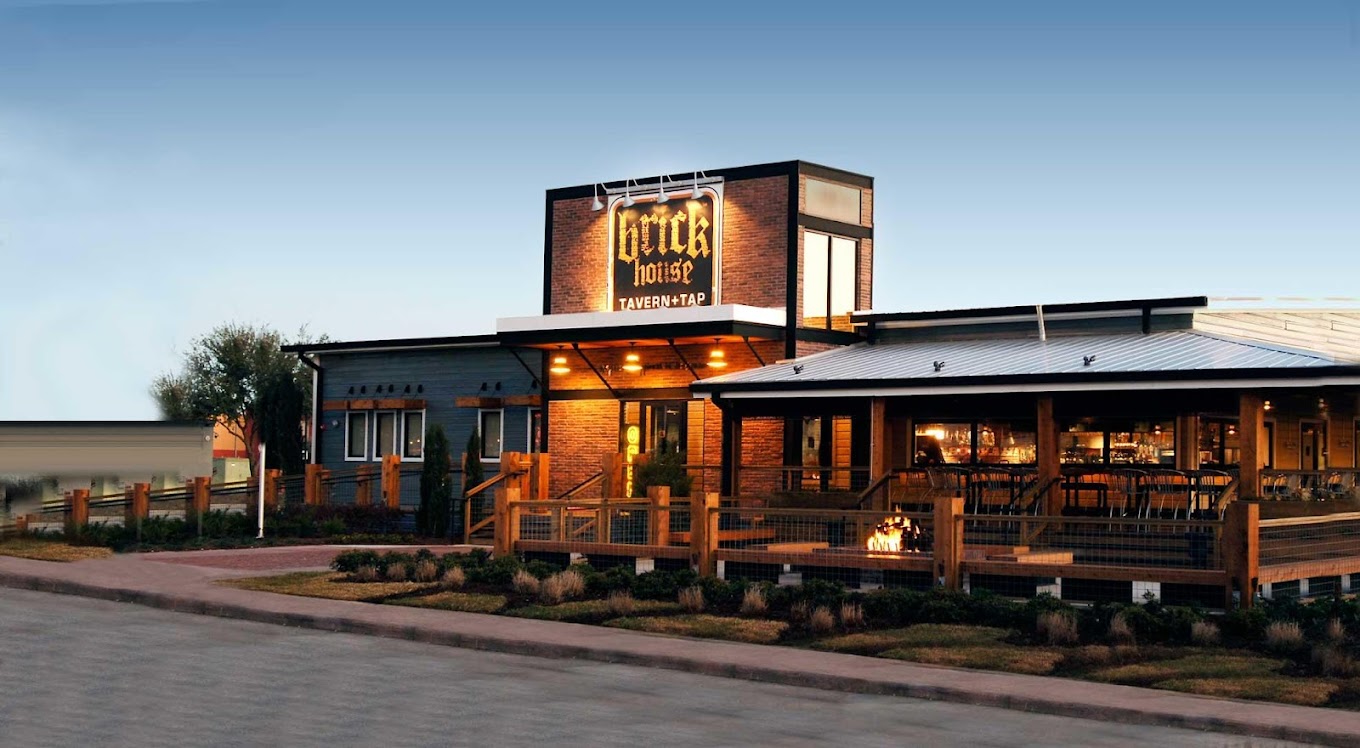 Restaurant/Tavern with brick facade and outdoor dining area, warm lights, and a central fire pit surrounded by a wooden fence at dusk.
