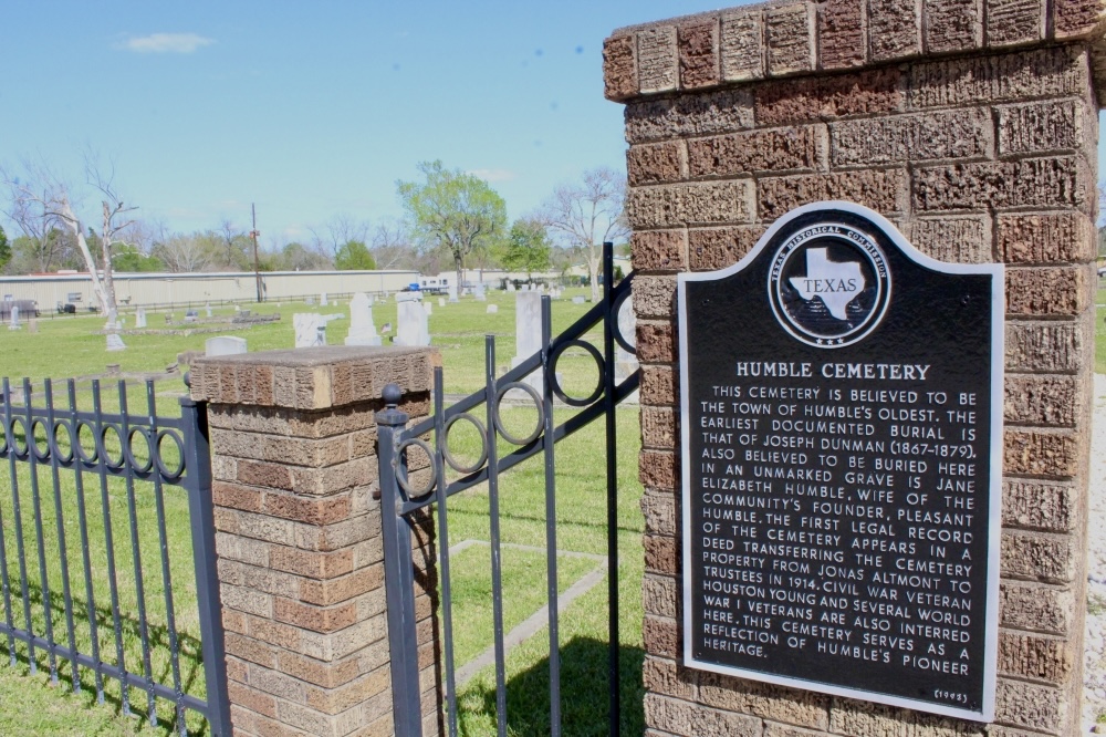 Brick entrance pillar with a black iron gate and a plaque for Humble Cemetery, graves visible in the background under a blue sky in Texas.