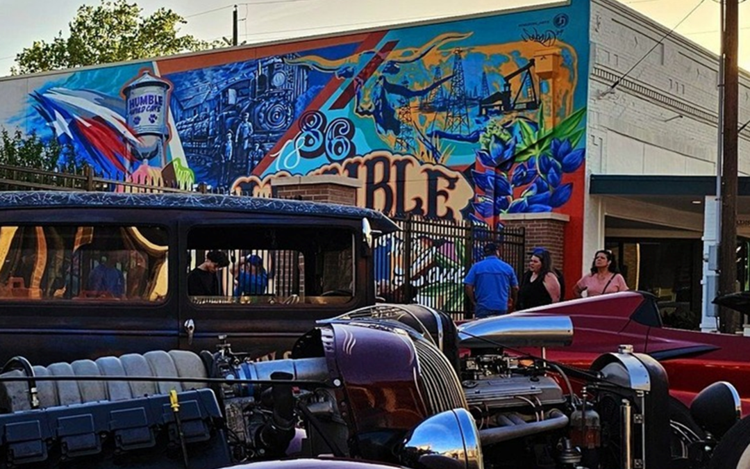 Vintage cars parked along a street with a vibrant mural on a building in the background, people walking by during a car show at sunset.